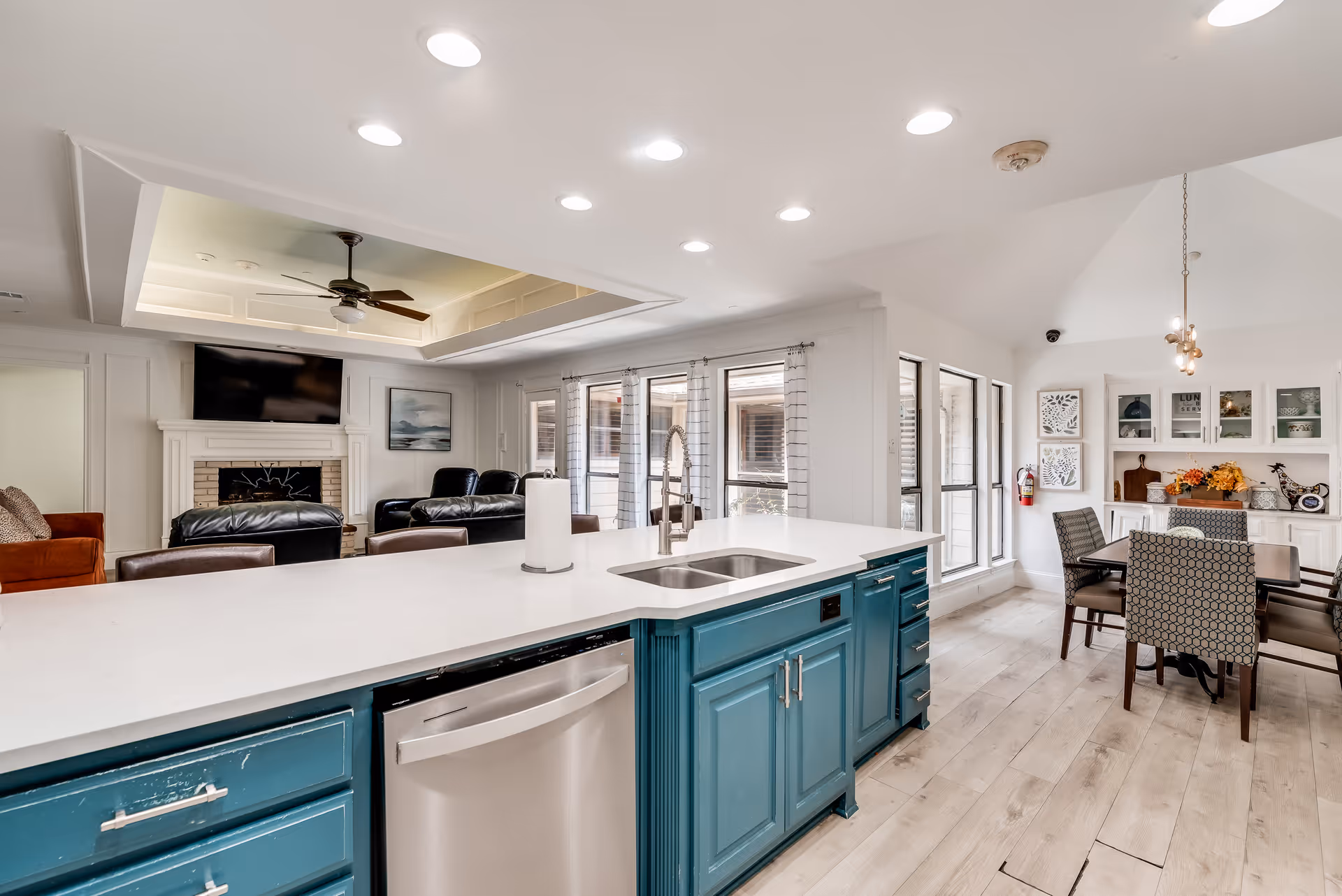 Bright and spacious open-concept living area featuring a kitchen island with a sink and dishwasher, teal cabinetry, a dining table with chairs, large windows with curtains, and a living room with leather sofas, a ceiling fan, and a wall-mounted TV above a fireplace.