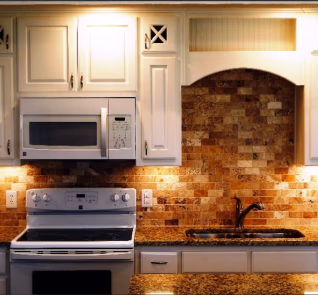 A kitchen interior featuring white cabinetry, a built-in microwave above a white electric stove, a granite countertop, a stainless steel sink with a faucet, and a brick-patterned backsplash with warm lighting underneath the cabinets.