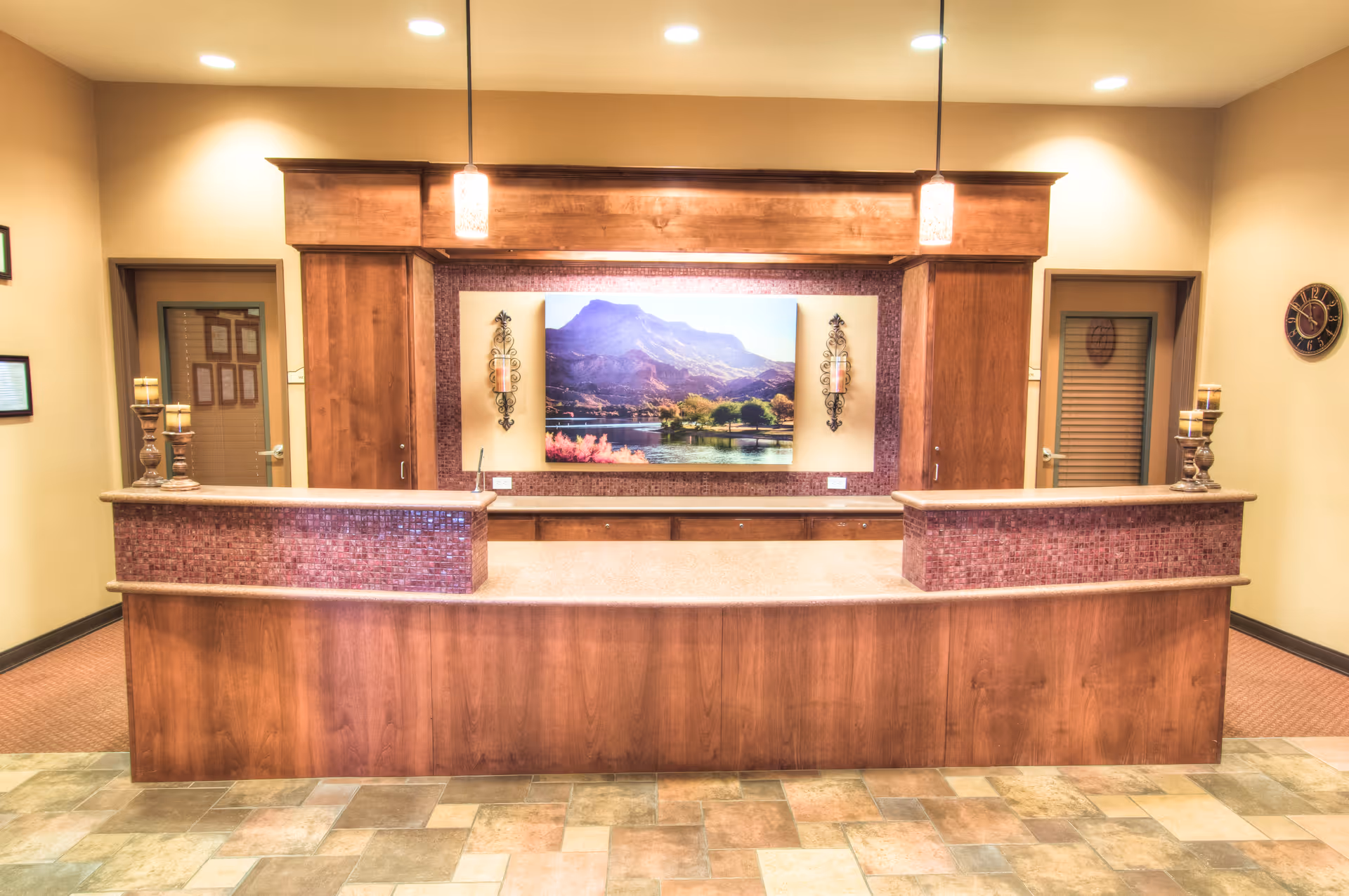 Reception desk area with wooden cabinetry and a countertop, two pendant lights hanging from the ceiling, a large landscape painting of mountains and a lake on the wall behind the desk, two doors on either side, and decorative candle holders on the counter.
