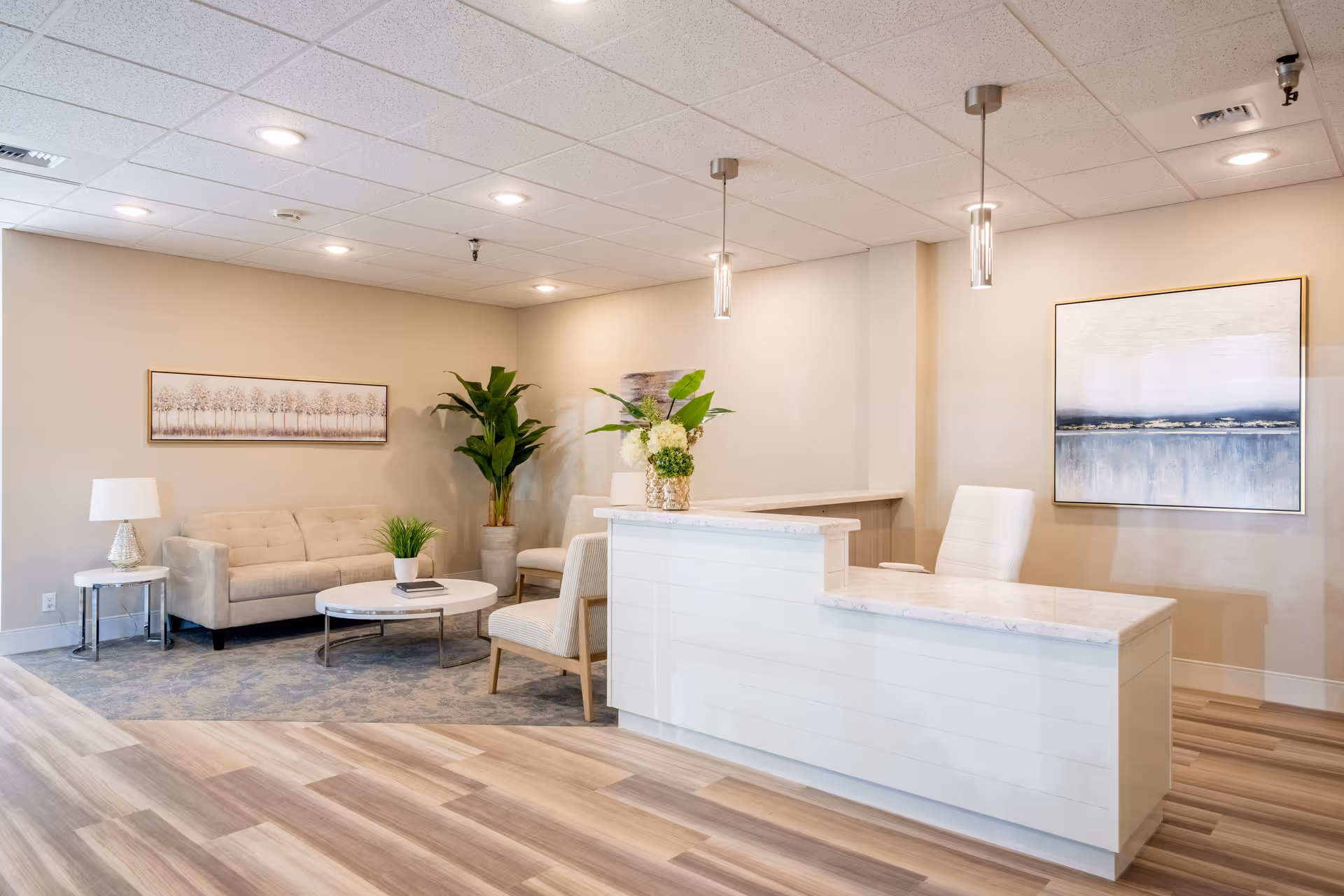 A bright and modern senior living facility reception area with a white reception desk, a white office chair, two pendant lights hanging from the ceiling, a beige sofa, two beige armchairs, a round white coffee table with a small plant and book, a side table with a lamp, two large framed artworks on the walls, and a tall potted plant in the corner. The floor is a combination of wood and carpet.
