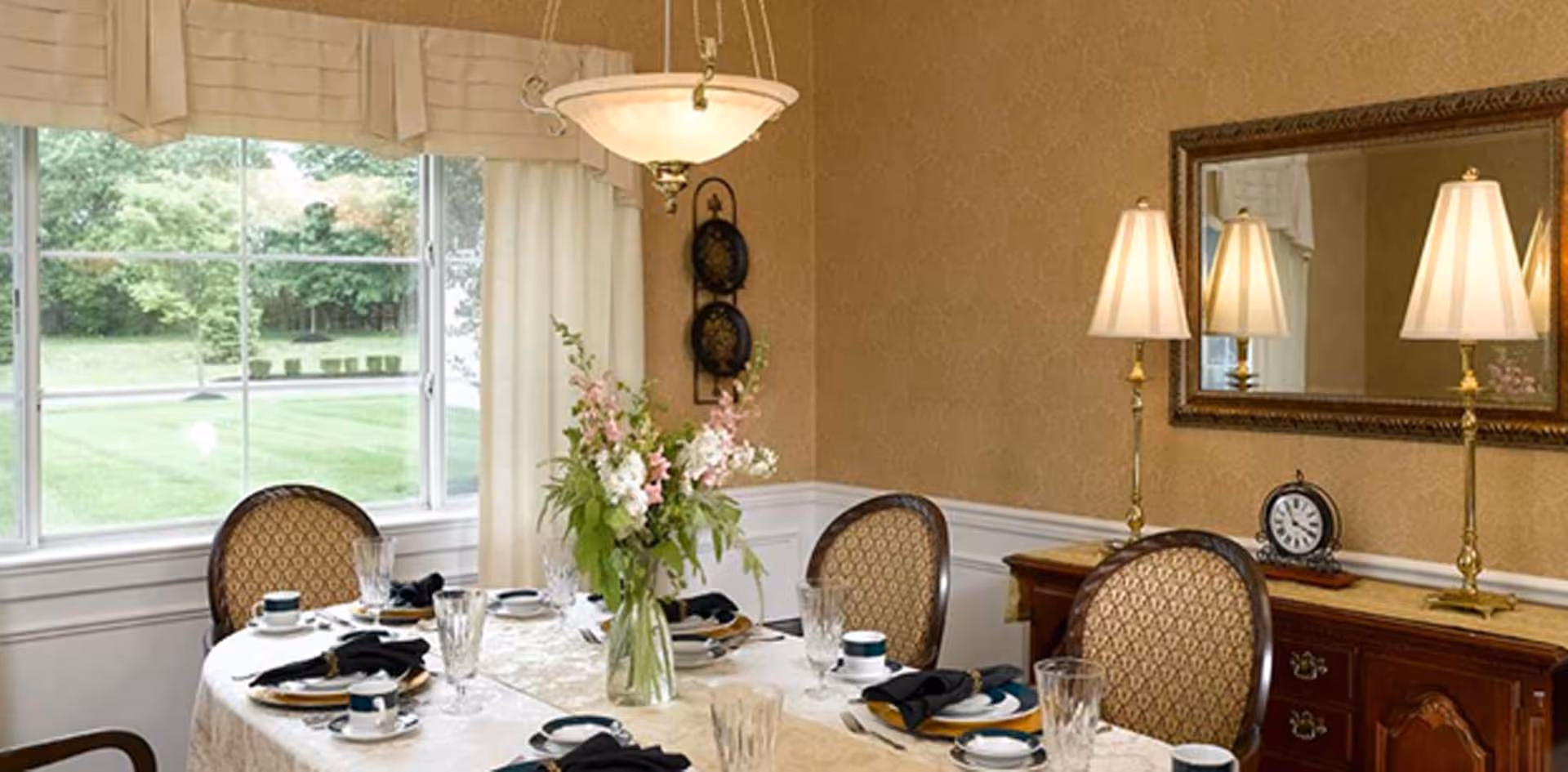 A formal dining room with a round table set for six people. The table is covered with a white tablecloth and has a vase with pink and white flowers as a centerpiece. Each place setting includes a plate, cup, saucer, glass, and black napkin. The room has beige wallpaper, a large window with cream curtains, a wooden sideboard with two lamps and a clock, and a large mirror above the sideboard.
