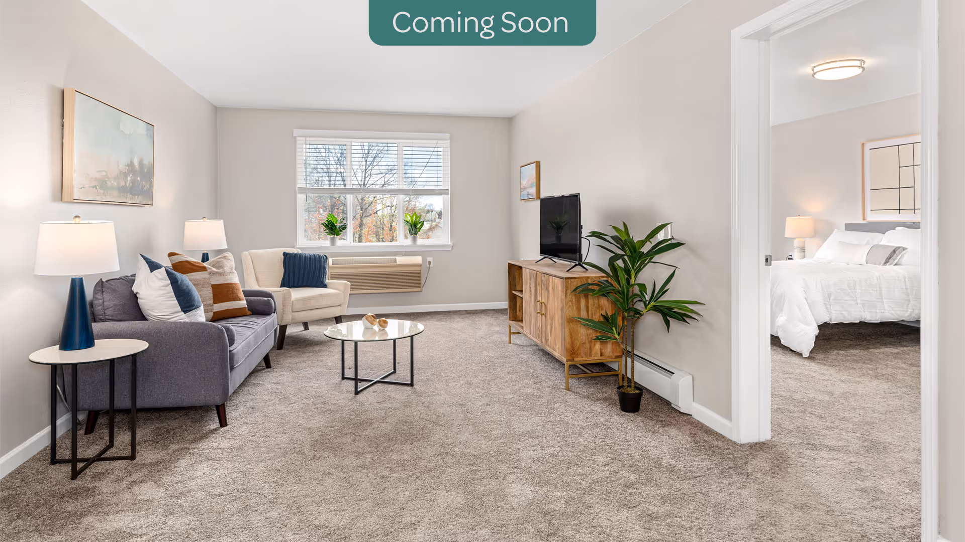 A bright and clean living room with beige walls and carpeted floor. The room features a gray sofa with multiple cushions, a beige armchair, a round glass coffee table, and two side tables with lamps. A wooden TV stand holds a flat-screen television, and a potted plant is placed beside it. A window with white blinds lets in natural light. Through an open doorway, a bedroom with a white bed and bedside lamp is visible.