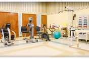 A rehabilitation exercise room with various fitness equipment including stationary bikes, an exercise ball, and other physical therapy machines. The room has a clean, bright interior with beige and white walls and a patterned border near the ceiling.