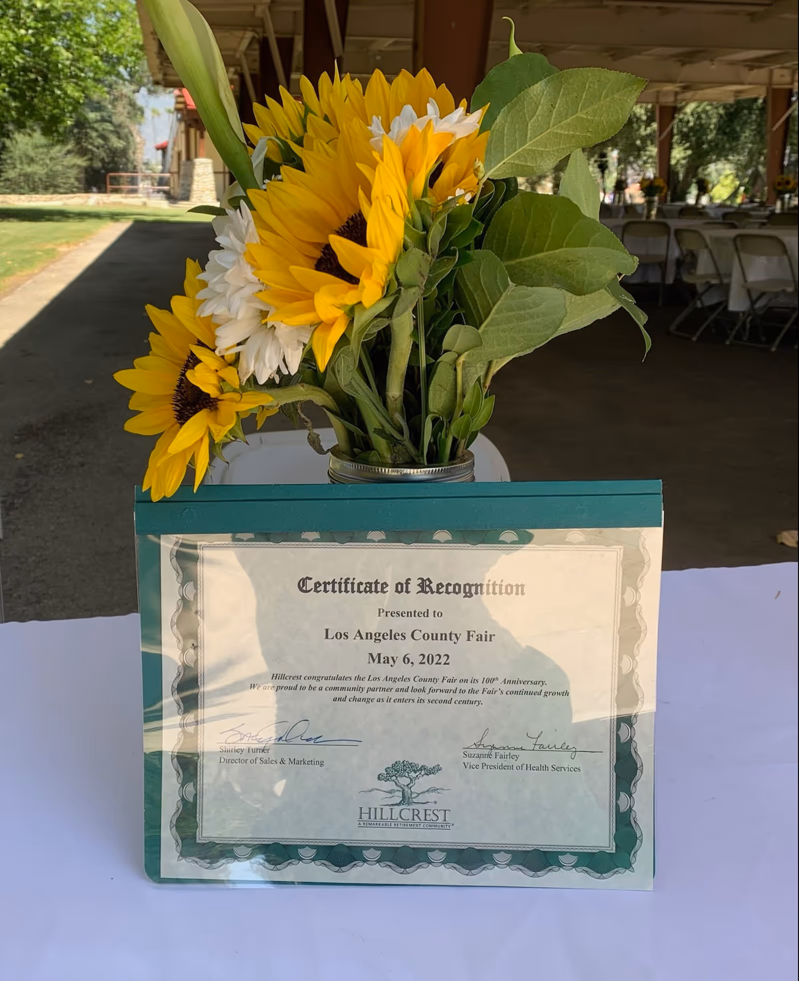A certificate of recognition presented to the Los Angeles County Fair on May 6, 2022, displayed on a table with a bouquet of sunflowers and white flowers in a jar behind it. The setting appears to be an outdoor covered area with tables and chairs in the background.
