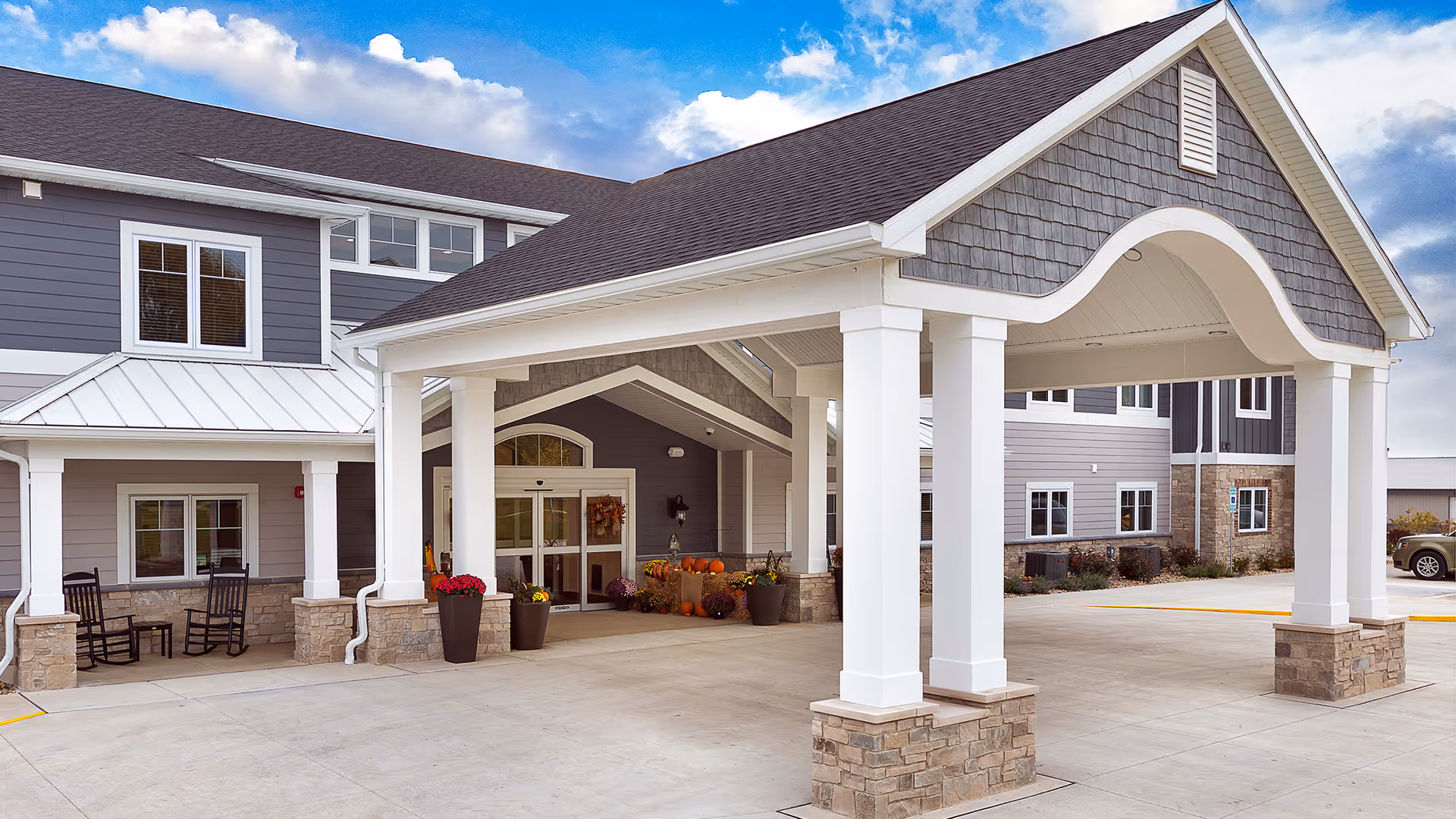 Entrance of Villas of Holly Brook Assisted Living & Memory Care facility in Bloomington Fox Creek, IL, featuring a covered drop-off area with white columns and stone bases, a gray building exterior with white trim, potted plants, and rocking chairs near the entrance under a partly cloudy sky.