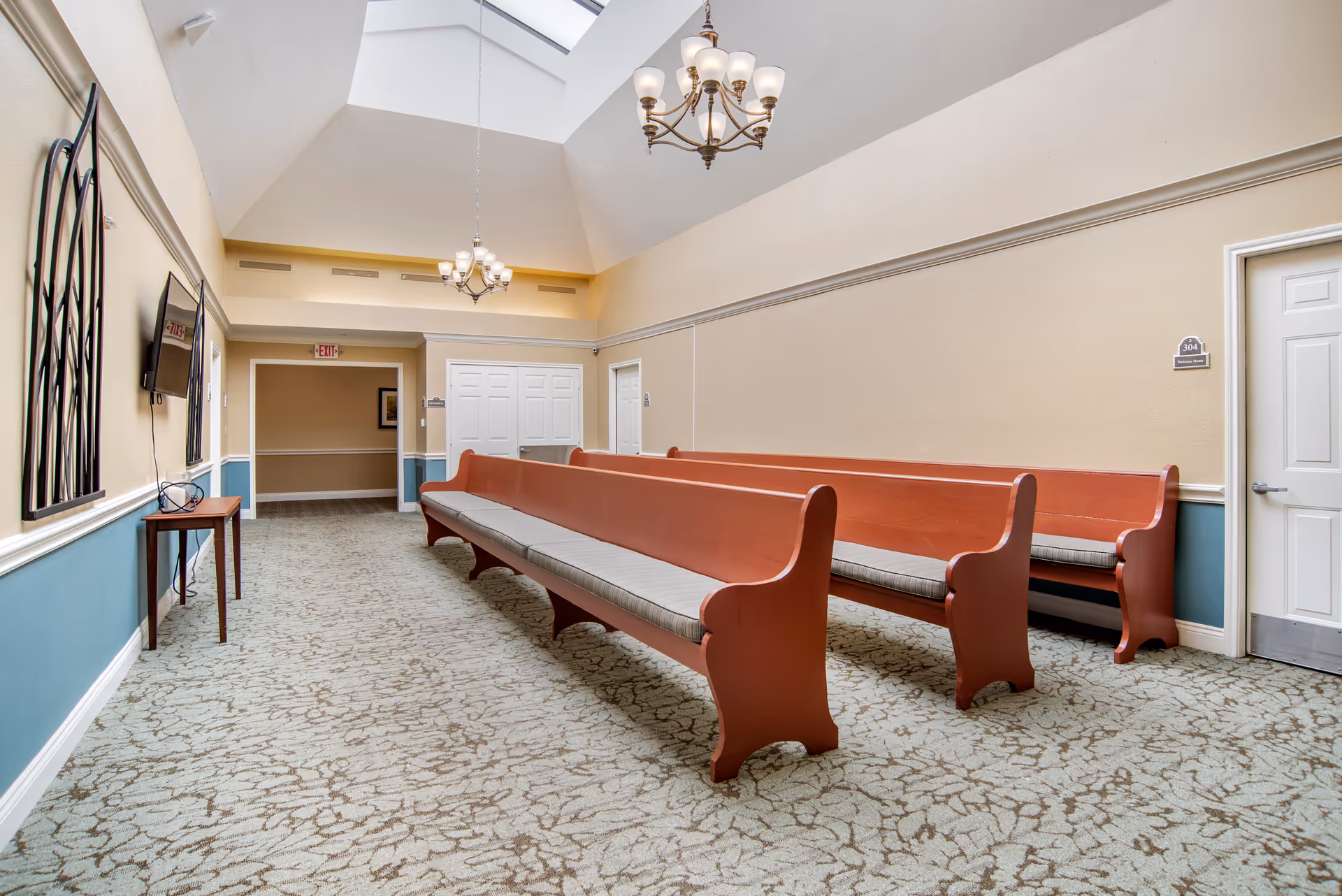 Interior view of a waiting area with long wooden benches with cushions, beige walls with blue wainscoting, carpeted floor, two chandeliers hanging from a high ceiling with skylights, a small table with a TV mounted on the wall, and several doors including one labeled 304.
