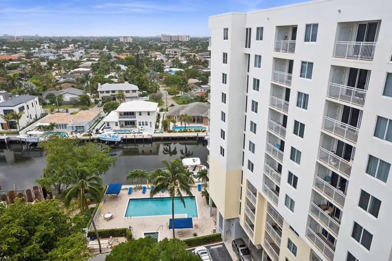 View of a multi-story residential building next to a canal with boats docked, a swimming pool with lounge chairs and palm trees in the foreground, and a neighborhood with houses and greenery in the background under a clear sky.