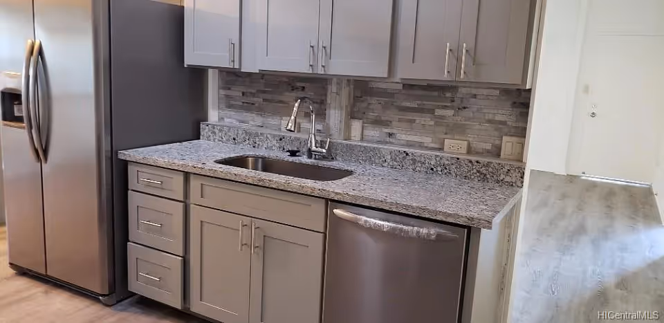 Modern kitchen area with stainless steel refrigerator, dishwasher, and a sink set in a granite countertop. The backsplash features a stone tile design, and there are light gray cabinets above and below the counter. The floor is light-colored wood, and a white door is visible in the background.
