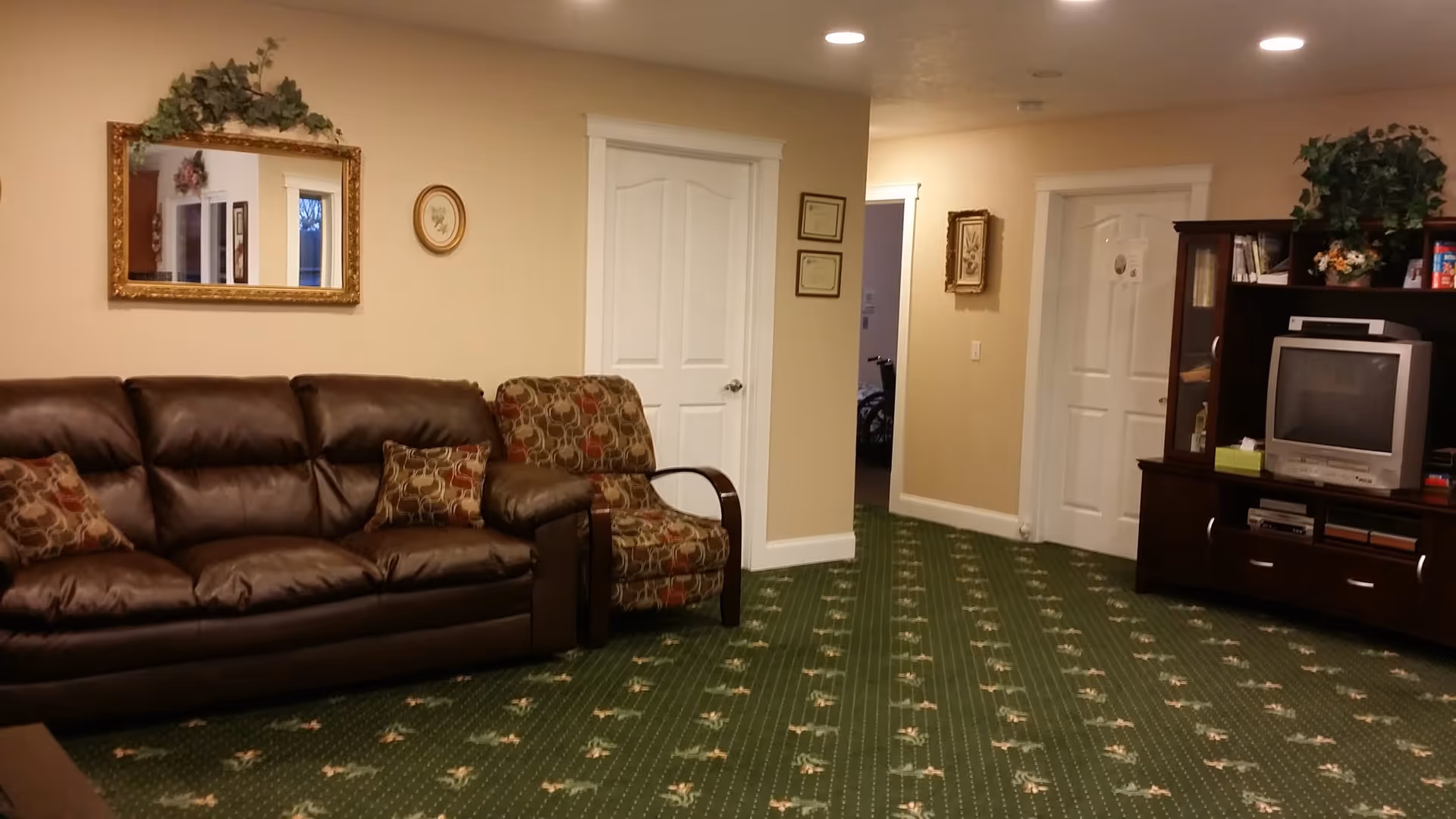 A cozy living room area with a brown leather sofa and a patterned armchair on a green carpet with floral designs. There is a large ornate mirror with greenery above it on the beige wall, two white doors, framed pictures, and a dark wooden entertainment center with an older-style television and decorative plants.