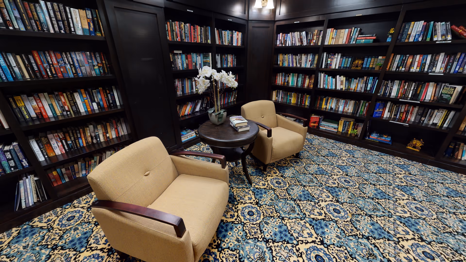 A cozy library room with dark wooden bookshelves filled with books lining the walls. Two beige armchairs with wooden armrests are positioned around a small round wooden table that holds a vase with white flowers and a few books. The floor is covered with a patterned blue and beige carpet.