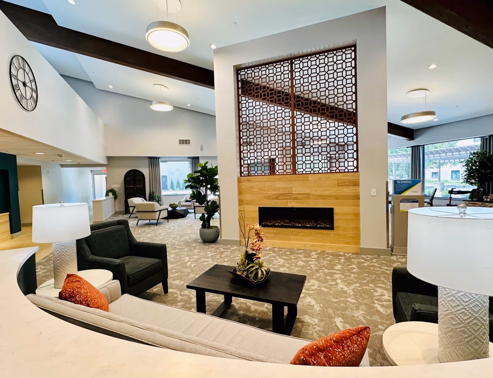 A spacious and modern living room area in Westminster Village featuring comfortable seating with armchairs and a sofa adorned with orange pillows. The room has a patterned carpet, a black coffee table with a decorative plant, and a large decorative partition with a geometric design above a wooden panel with an electric fireplace. Large windows allow natural light to fill the space, and there are ceiling lights and a large wall clock visible.