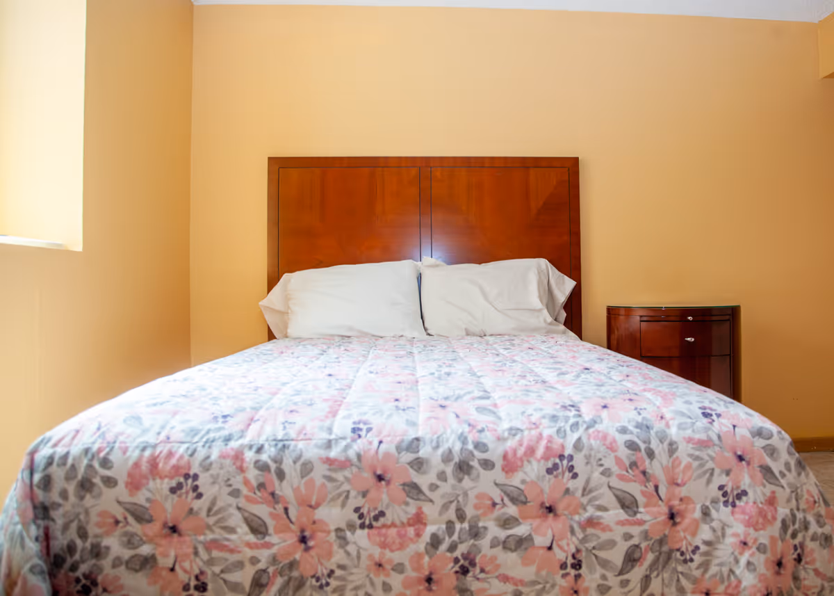A neatly made bed with a floral bedspread, wooden headboard, two pillows, and a small wooden nightstand in a peach-colored bedroom.