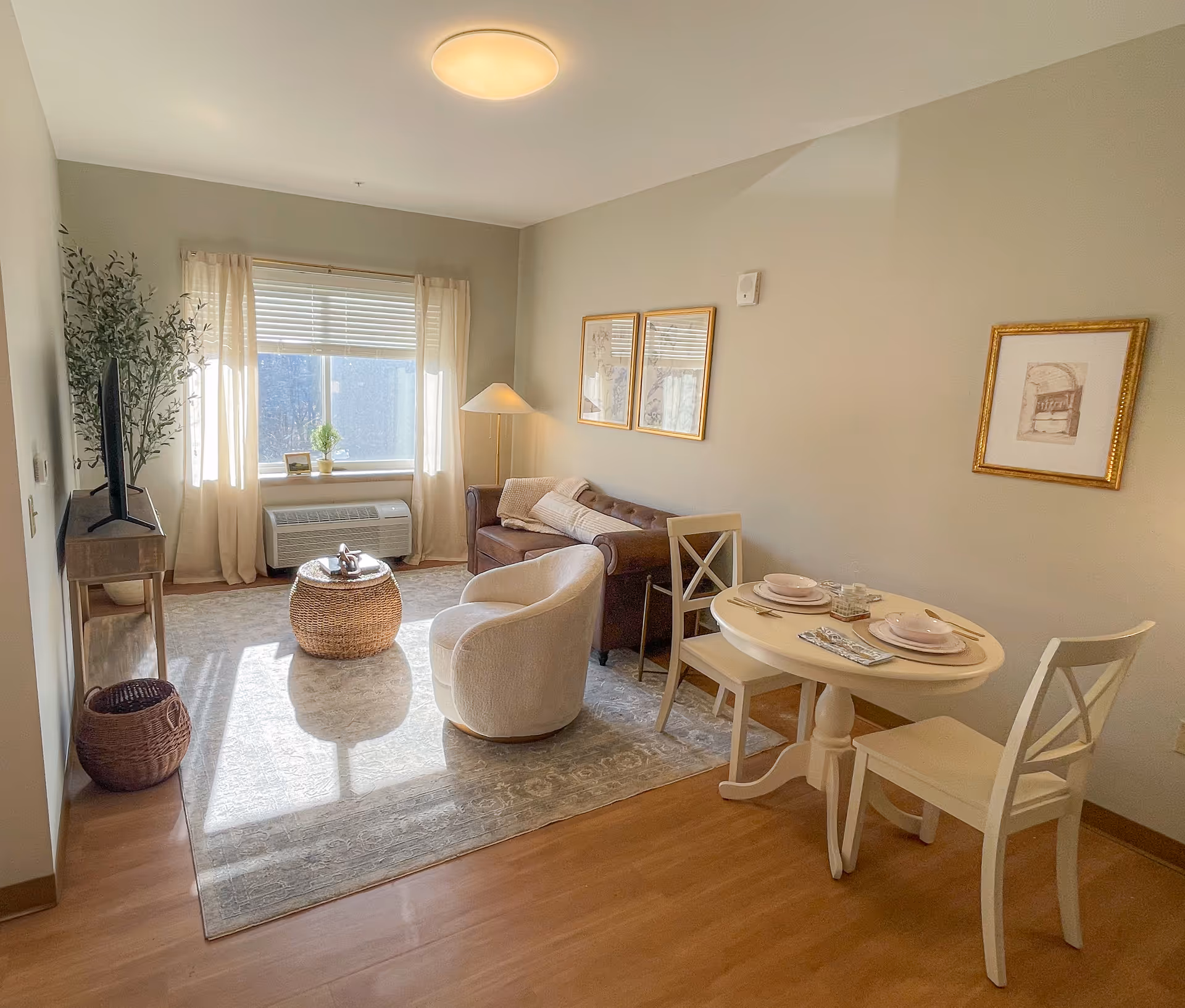 Sunlit living room with a sofa, armchair, TV, small round dining table and a window with curtains.