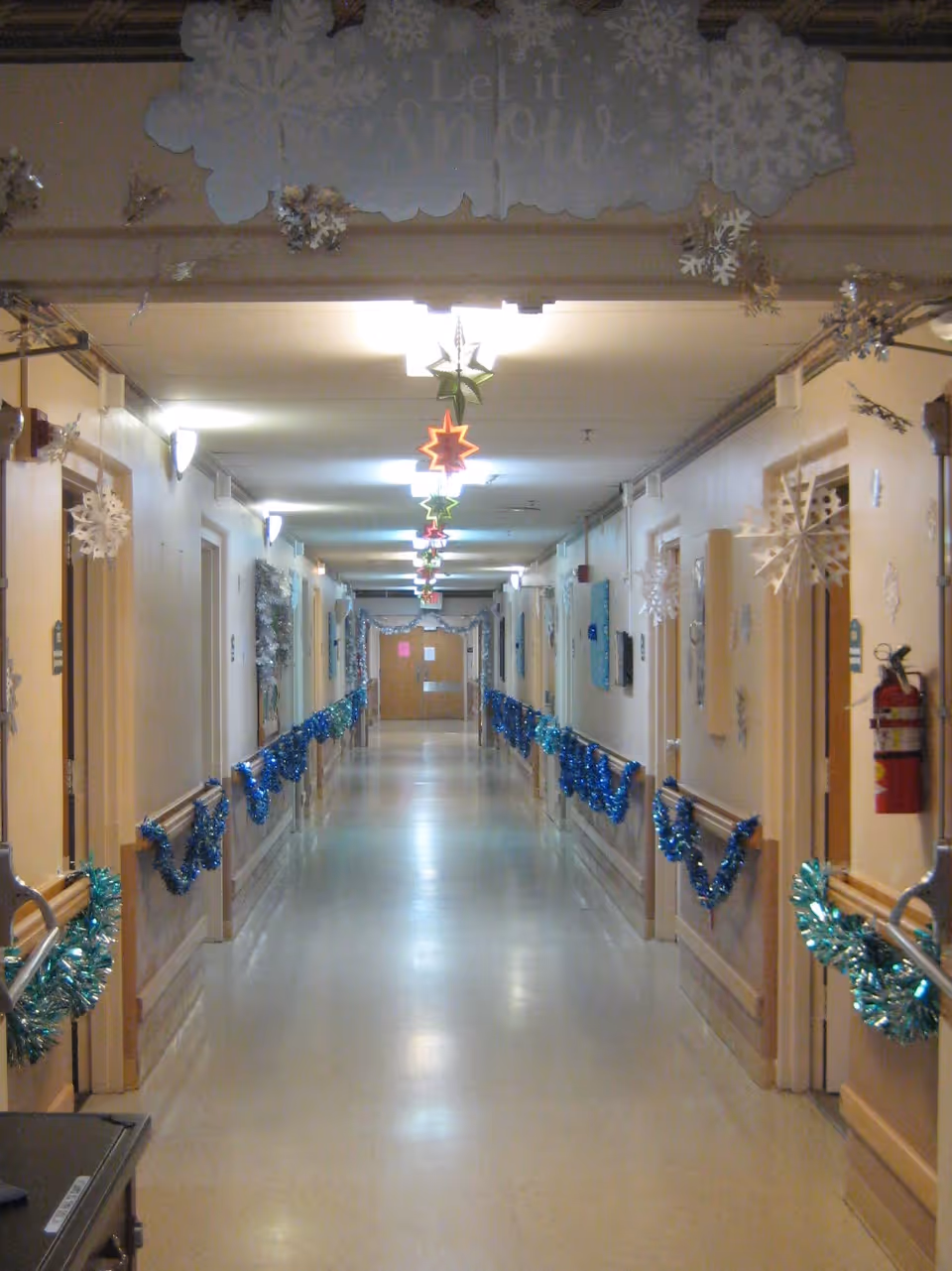 A long hallway in a care facility decorated with blue tinsel garlands along the walls and hanging snowflake and star decorations from the ceiling. The hallway has beige walls, handrails, and several doors on both sides. A sign at the top reads 'Let it Snow'.