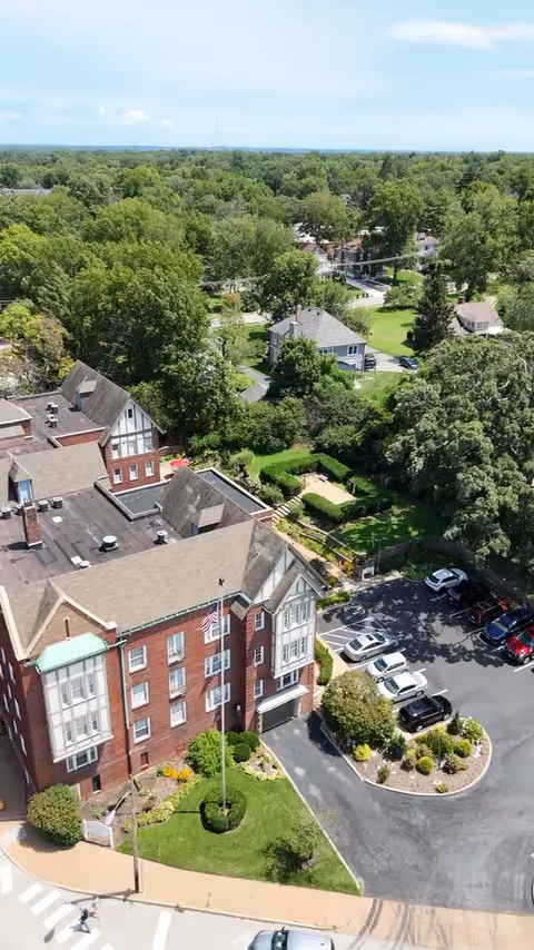 Aerial view of a large brick building with multiple windows and a flagpole in front, surrounded by greenery and trees. There is a parking lot with several cars parked, and residential houses and more trees are visible in the background under a clear blue sky.
