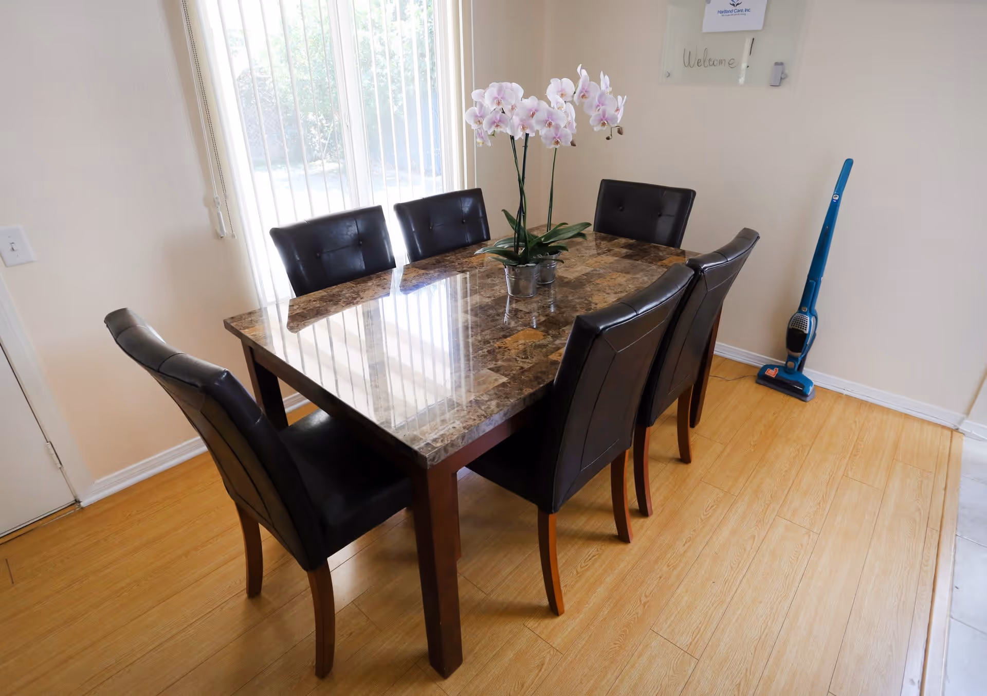 A dining area with a rectangular marble-top table surrounded by six black cushioned chairs. A potted orchid plant is placed in the center of the table. There is a large window with vertical blinds letting in natural light, and a blue vacuum cleaner is standing against the wall on the right side.