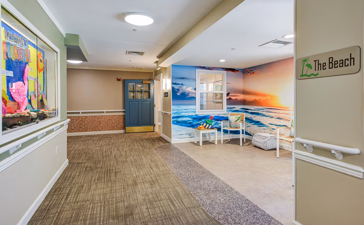 Interior hallway of a memory care community with a seating area named 'The Beach' featuring a large wall mural of a beach sunset, two white chairs with colorful cushions, a small table with towels, and a wicker basket. The hallway has carpeted flooring, beige walls with handrails, and a blue door at the end. A bulletin board with colorful decorations is visible on the left wall.
