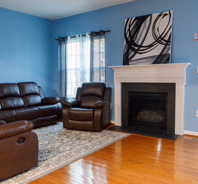 A cozy living room with blue walls, a large window with sheer curtains, a white fireplace with a modern black and white abstract painting above it, and three brown leather recliner chairs arranged on a patterned area rug over a polished wooden floor.