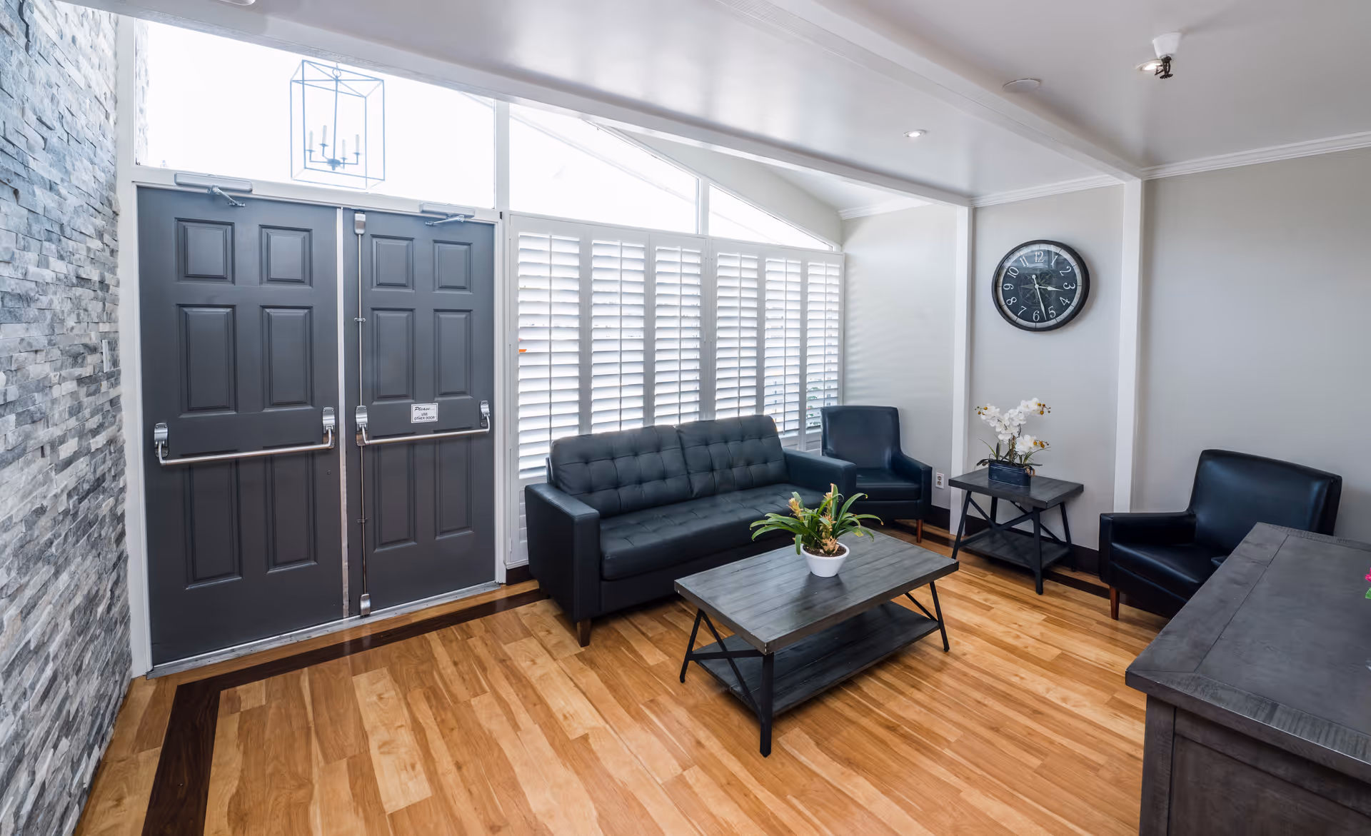 A bright and modern living room area with a black sofa, two black armchairs, a wooden coffee table with a potted plant, and a side table with white flowers. The room has light wooden flooring, a stone accent wall, large windows with white shutters, and a double gray door with a sign. A round black wall clock is mounted on the light gray wall.