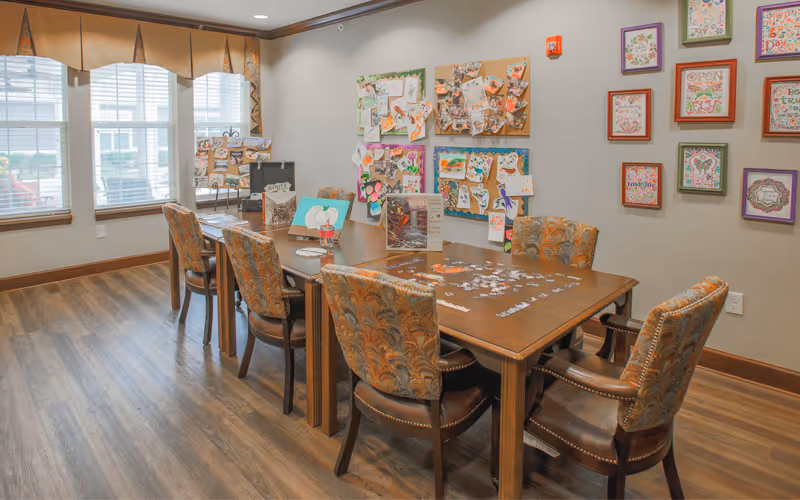 A well-lit activity room with a wooden table surrounded by six upholstered chairs. The table has a partially completed jigsaw puzzle and some art supplies on it. The walls are decorated with colorful artwork and bulletin boards displaying various crafts and notes. Large windows with valances let in natural light, and the floor is wood.