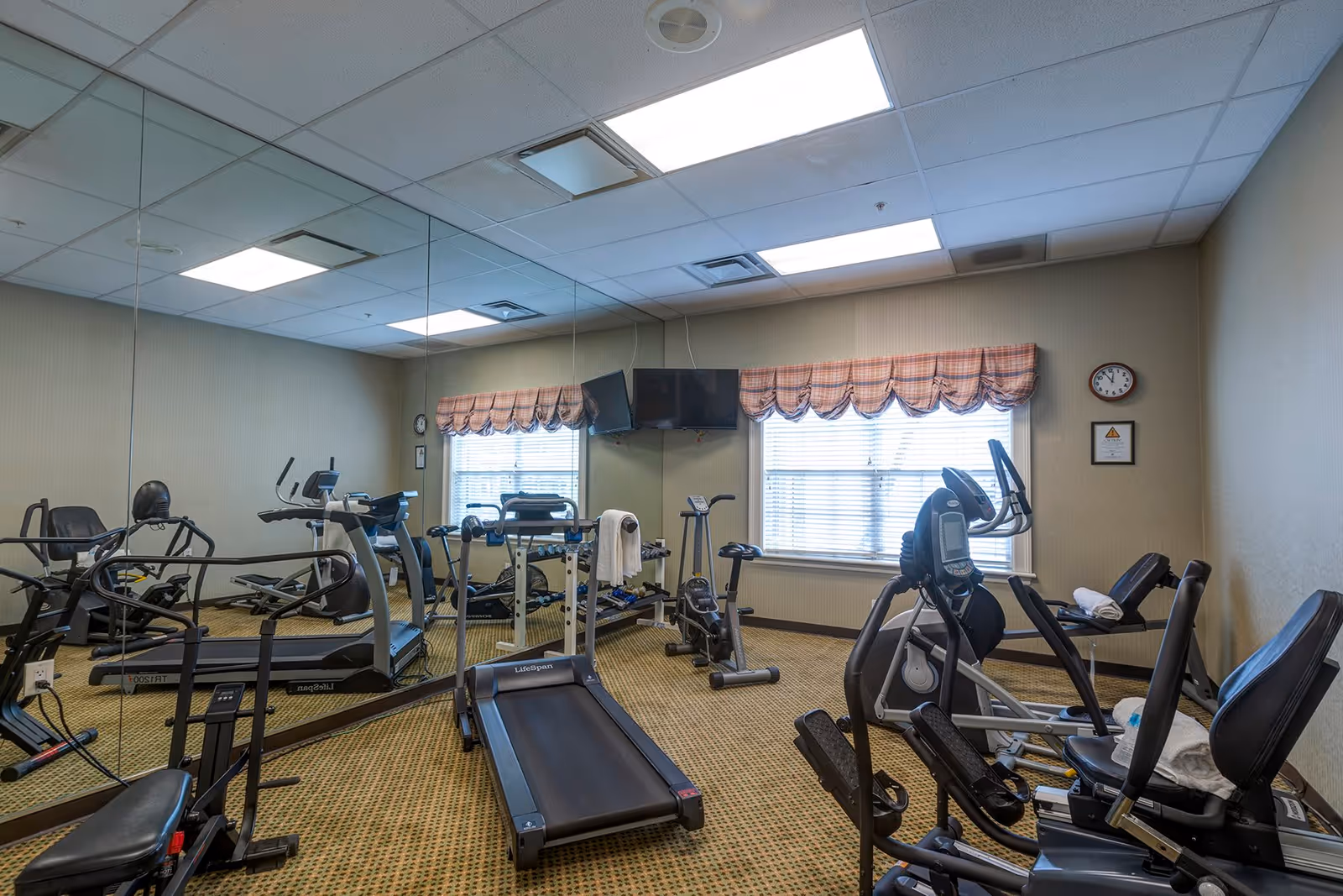 A small fitness room with various exercise equipment including treadmills, stationary bikes, and weight machines. The room has a large mirror covering one wall, two windows with plaid valances, a wall clock, and a mounted TV.
