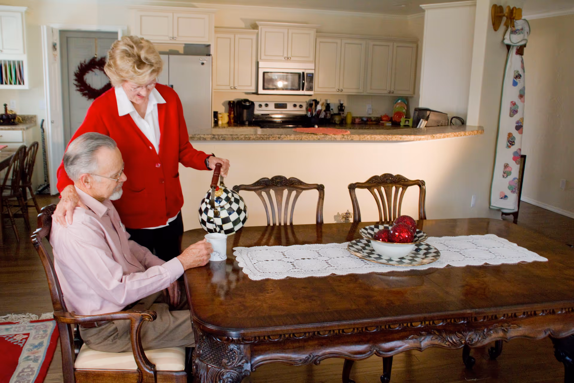 An elderly woman in a red cardigan is pouring a beverage from a checkered teapot into a cup held by an elderly man seated at a wooden dining table with a white lace runner and a decorative bowl with red ornaments. The background shows a kitchen with white cabinets and appliances.