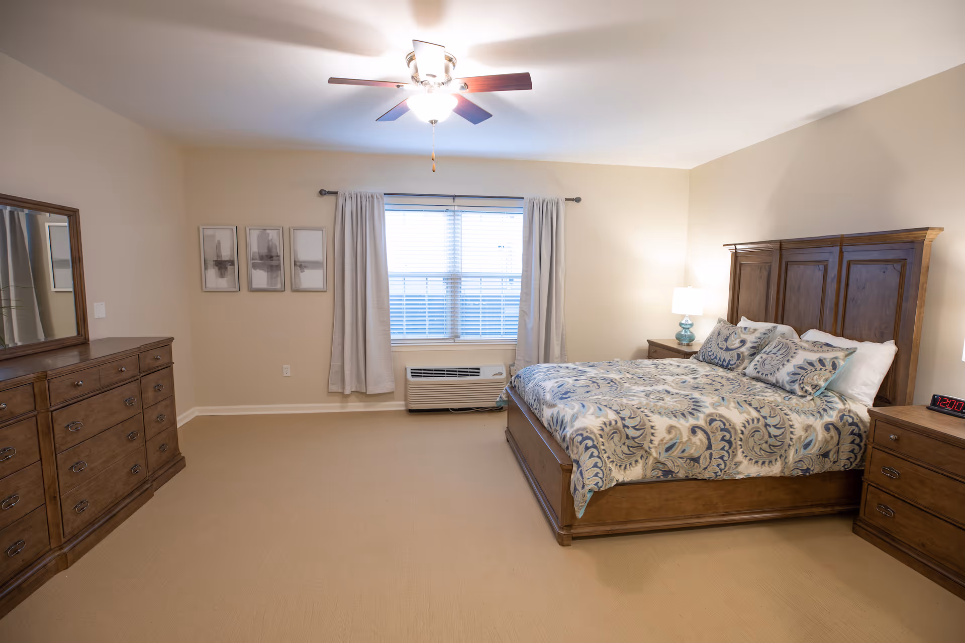 A well-lit bedroom with a large wooden bed featuring a patterned blue and beige bedspread and matching pillows. The room has beige walls and carpet, a ceiling fan with lights, a window with white blinds and light-colored curtains, a wooden dresser with a mirror on the left, and two wooden nightstands with lamps on either side of the bed.