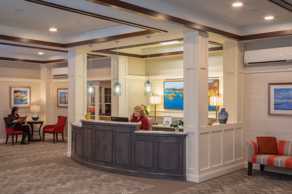 Interior view of a senior living facility reception area with a curved wooden front desk where a woman is talking on the phone. The space features beige walls, decorative columns, hanging pendant lights, framed landscape paintings, and comfortable seating including red chairs and a striped sofa with red cushions. Another woman is seated reading a magazine in the background.