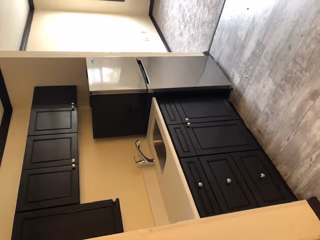 Small kitchen area with dark brown cabinets, a stainless steel sink, and a silver refrigerator. The kitchen has light-colored countertops and wood-patterned flooring. Adjacent to the kitchen is a carpeted room with beige walls and dark trim.