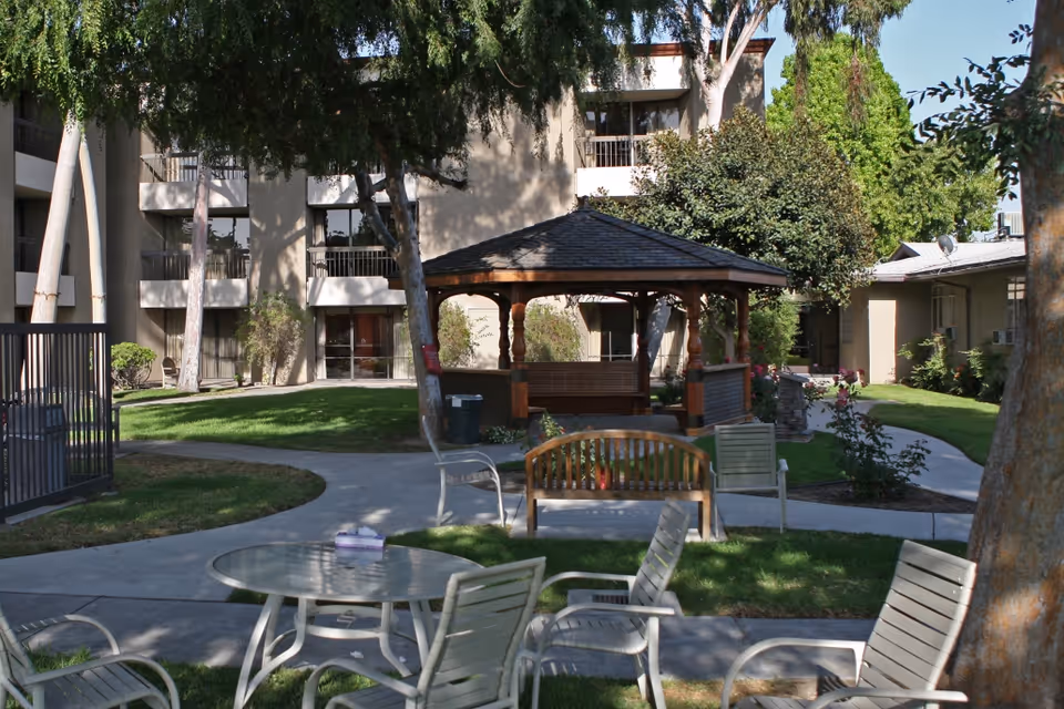 Sunlit courtyard with a wooden gazebo, benches, patio tables and chairs in front of a multi-story senior living building.