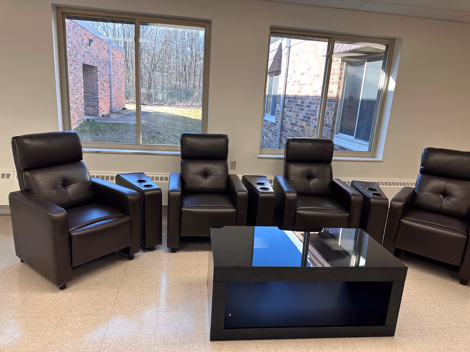 Four brown leather recliner chairs with side tables arranged around a black coffee table in a bright room with large windows looking out to a brick courtyard.