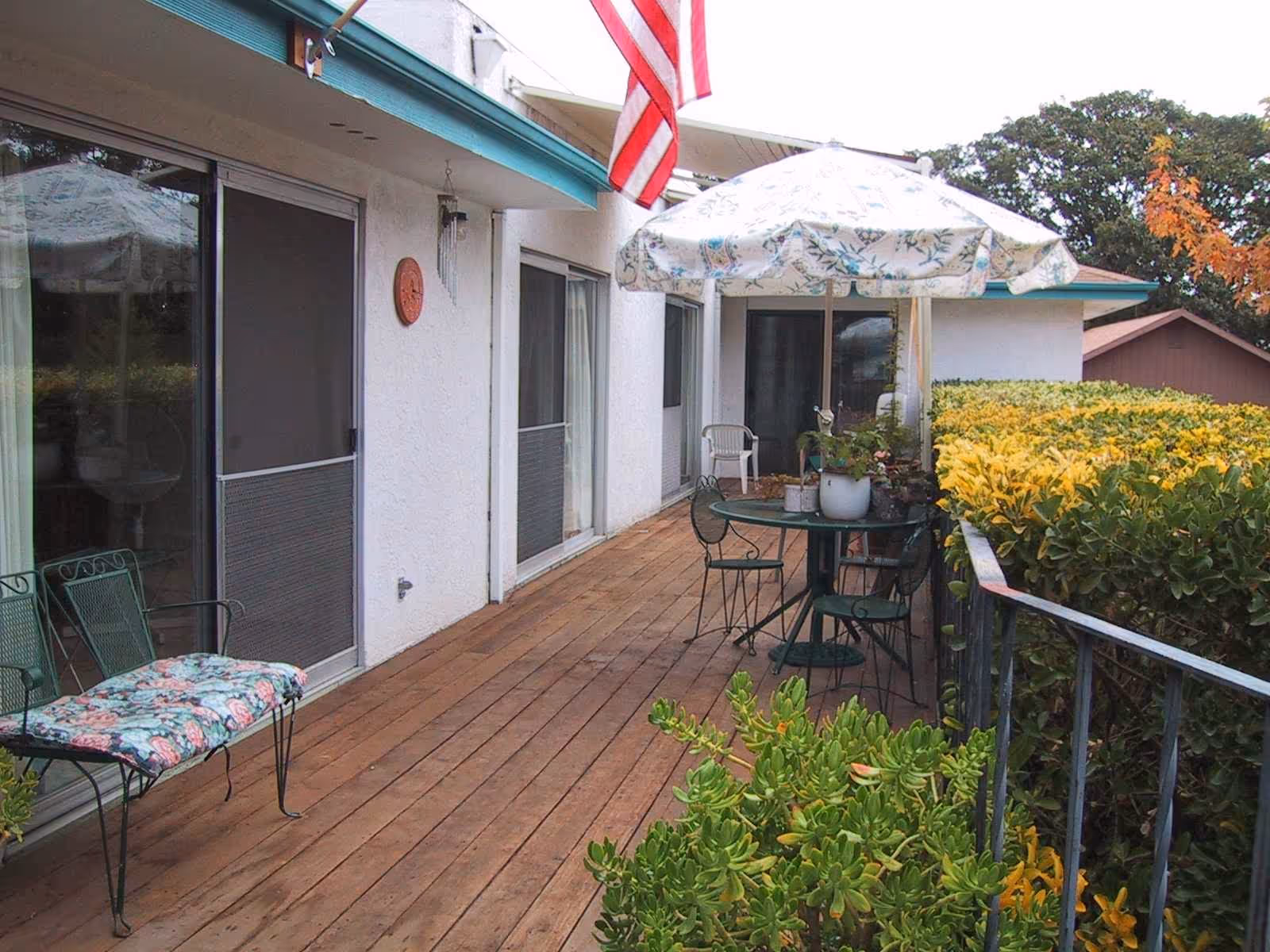 A wooden deck patio attached to a white building with sliding glass doors. The patio has a round table with a floral umbrella and several chairs. There is a bench with a floral cushion, potted plants on the table, and a hedge along the railing. An American flag is hanging above the deck.