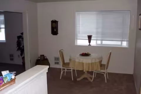 A small dining area with a round table covered with a beige tablecloth and two chairs. There is a window with closed blinds behind the table and a decorative vase on the windowsill. A wall clock is mounted on the wall to the left, and part of a plant is visible in an adjacent room.