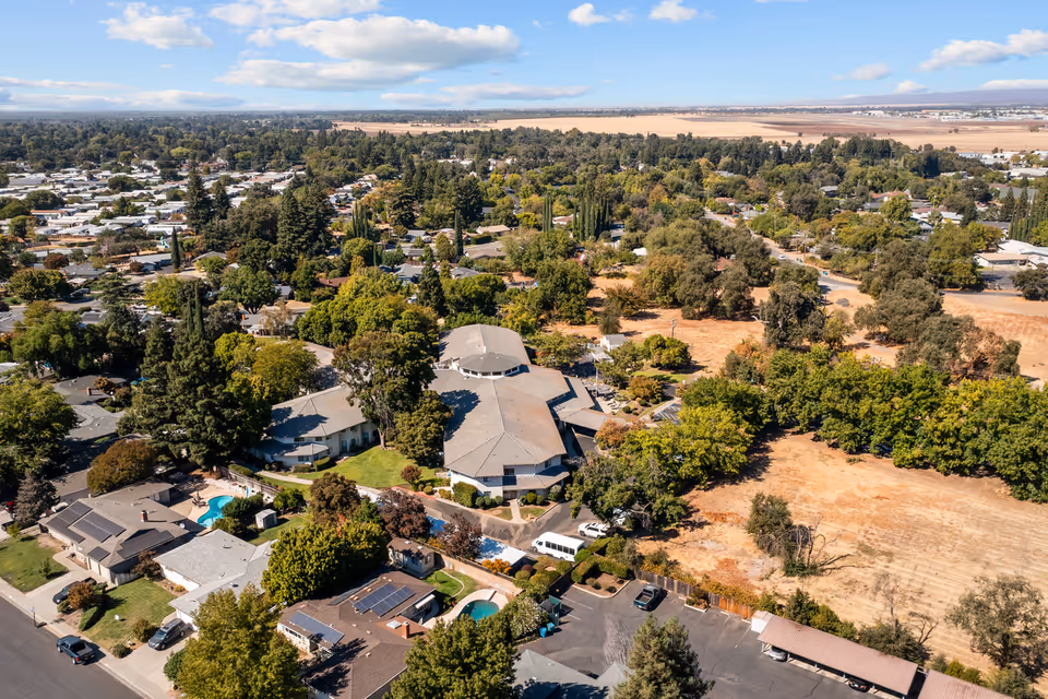Aerial view of a senior living facility named Provincial Chico Senior Living surrounded by trees, residential houses, and open fields under a partly cloudy sky.