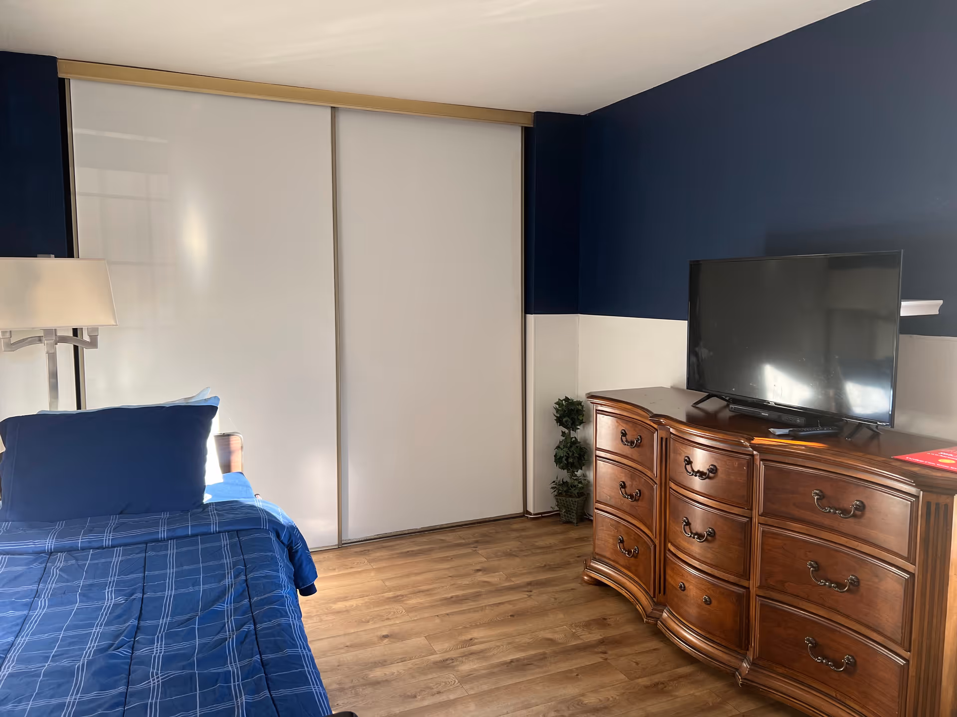 Bedroom with a blue-made bed, white sliding closet doors, and a wooden dresser topped by a TV.