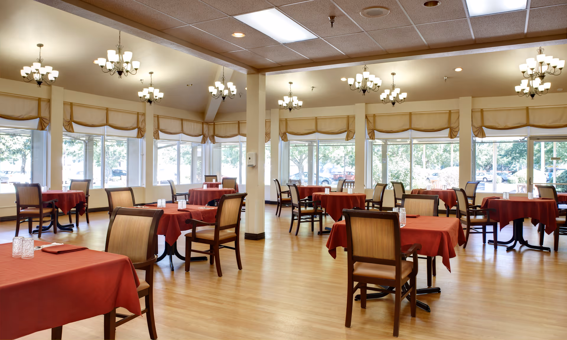 A spacious dining room with multiple tables covered in red tablecloths and surrounded by wooden chairs with cushioned seats. Large windows line the walls, allowing natural light to fill the room. Several chandeliers hang from the ceiling, providing additional lighting. The floor is made of light-colored wood.