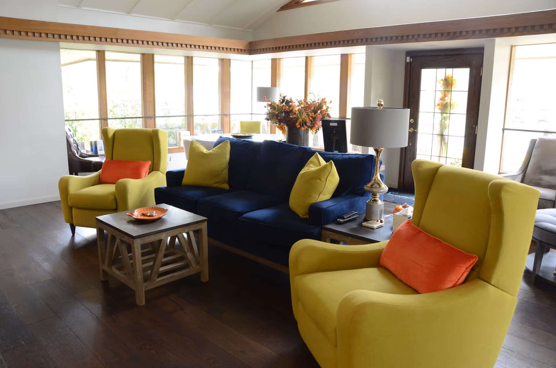 A bright living room with large windows letting in natural light. The room features a dark blue sofa with yellow pillows, two yellow armchairs with orange cushions, a wooden coffee table with an orange decorative plate, and a side table with a lamp and remote controls. There is a door with glass panels and a floral wreath, and a vase with autumn flowers on a table behind the sofa.