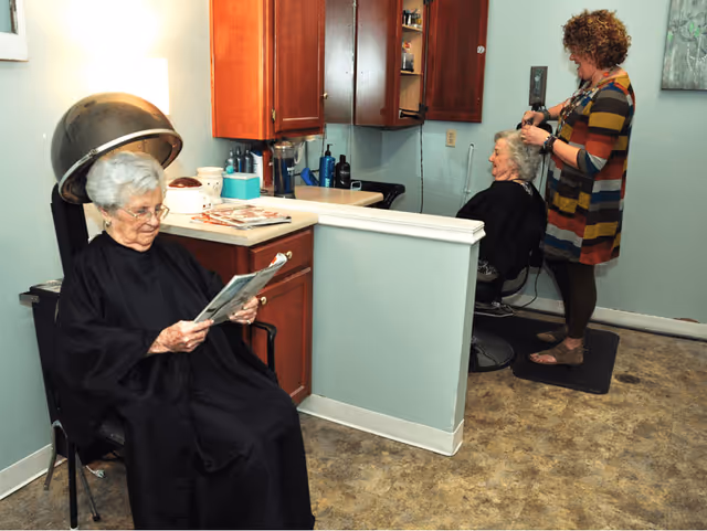 An elderly woman sits under a hair dryer reading a magazine while another elderly woman is getting her hair styled by a hairdresser in a small salon area with light blue walls and wooden cabinets.