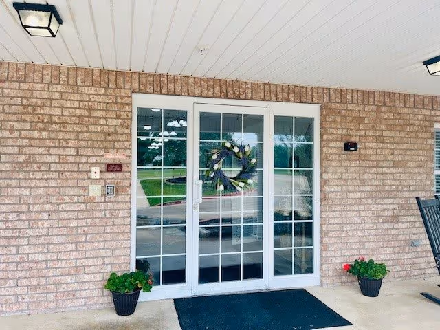Entrance to a building with a glass double door featuring a decorative wreath. The building has a brick exterior and a covered porch with two potted plants on either side of the door and a black doormat in front. There is a rocking chair partially visible on the right side.
