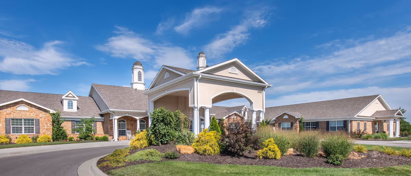 Front exterior of a single-story senior living facility with a covered entrance, circular driveway, and landscaped grounds under a blue sky.