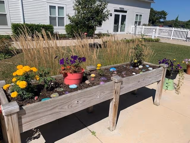 Raised wooden garden bed with various flowers and decorative painted stones, situated on a concrete patio in front of a white building with windows and a white picket fence in the background under a clear blue sky.