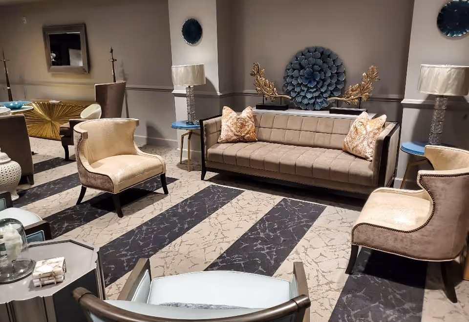 A stylish living room area with a beige sofa adorned with two decorative pillows, two cream-colored armchairs, and a patterned carpet with wide stripes. The room features two side tables with lamps, a decorative blue wall piece above the sofa, and a gold ornamental piece on a console table in the background.