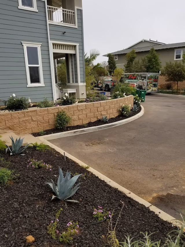 Exterior view of a residential building with blue siding and white trim, featuring a small porch area. In the foreground, there is a landscaped garden bed with various plants and mulch, bordered by a low retaining wall. A driveway curves around the garden bed, and in the background, there are other residential buildings and a vehicle with a gardening maintenance advertisement.