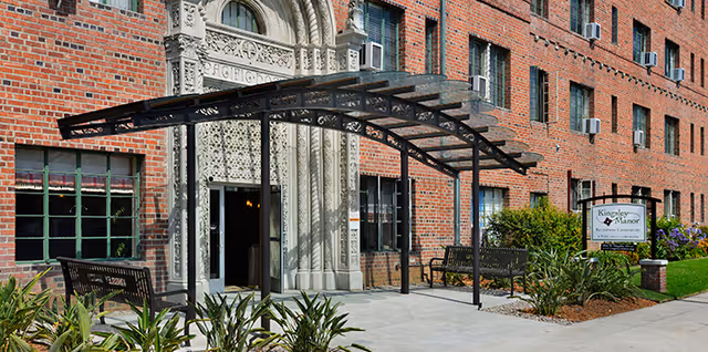 Entrance of a brick building with a decorative stone archway above the door, a black metal canopy, benches on either side, and a sign reading 'Kingsley Manor Assisted Living & Memory Care'. There are plants and greenery along the walkway leading to the entrance.