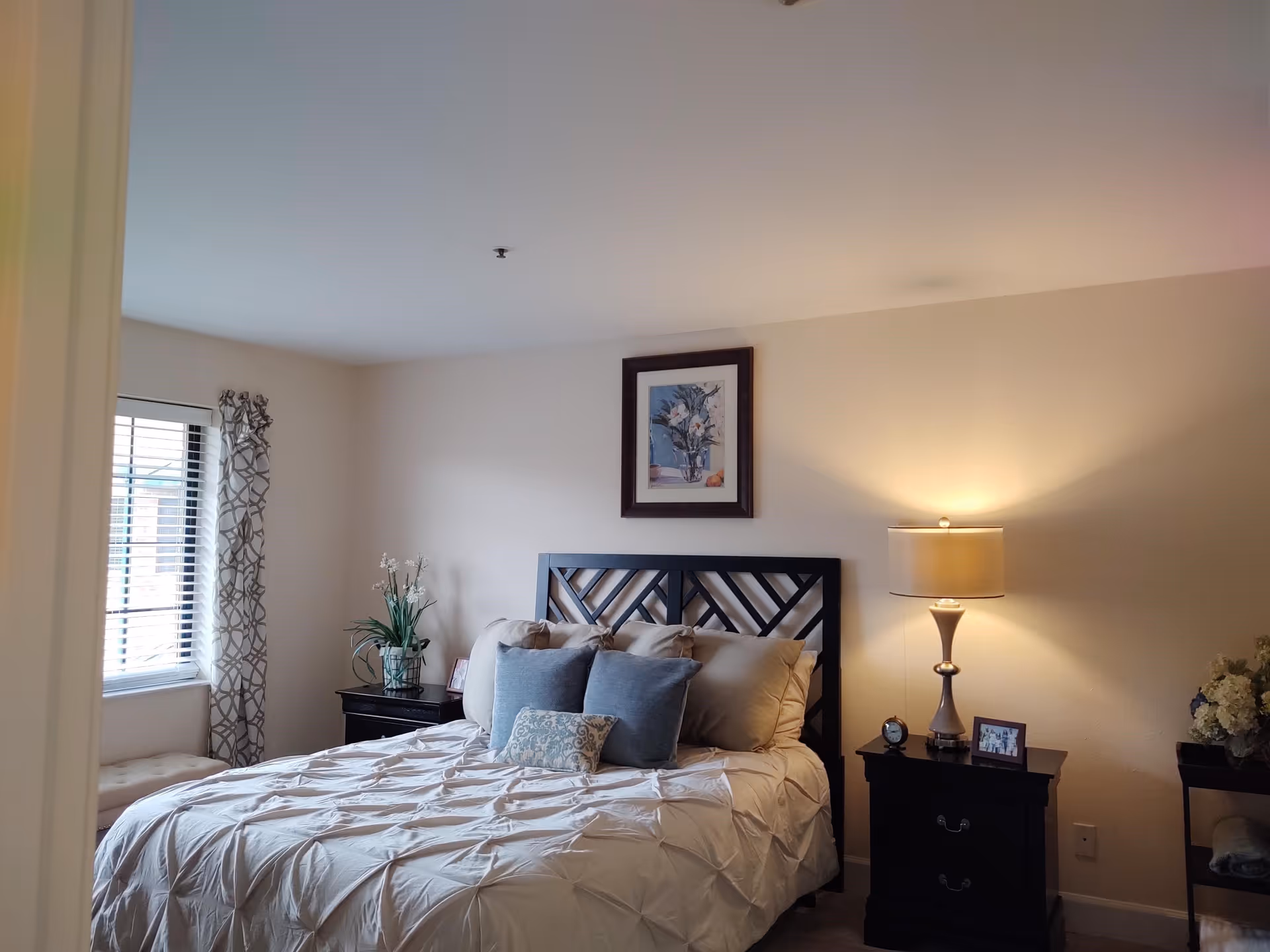 A cozy bedroom with a neatly made bed featuring beige and blue pillows. There are two dark wooden nightstands on either side of the bed, one with a lamp and a small clock, and the other with a plant and a picture frame. A window with patterned curtains is on the left side, and a framed floral artwork hangs above the bed.