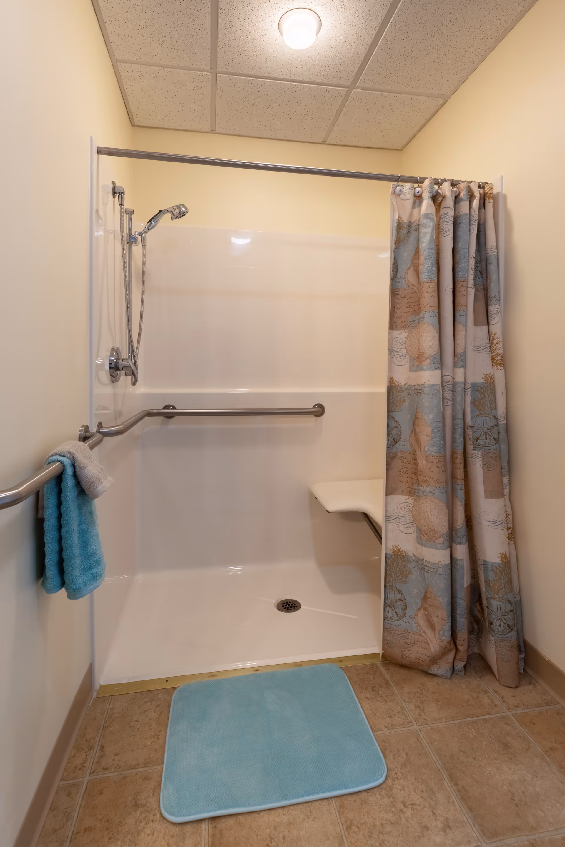 Accessible walk-in shower with grab bars, a fold-down seat, hand-held showerhead, patterned shower curtain and a blue bath mat.