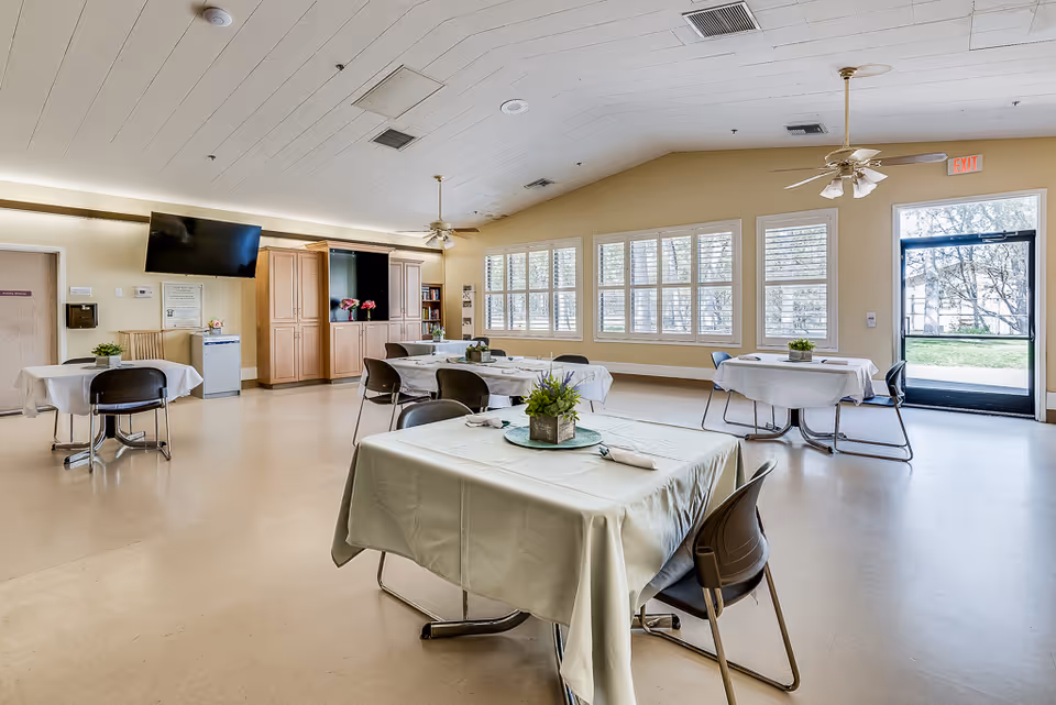 Spacious bright dining/activity room with several tables covered in tablecloths and centerpieces, large windows, ceiling fans and a wall-mounted TV.