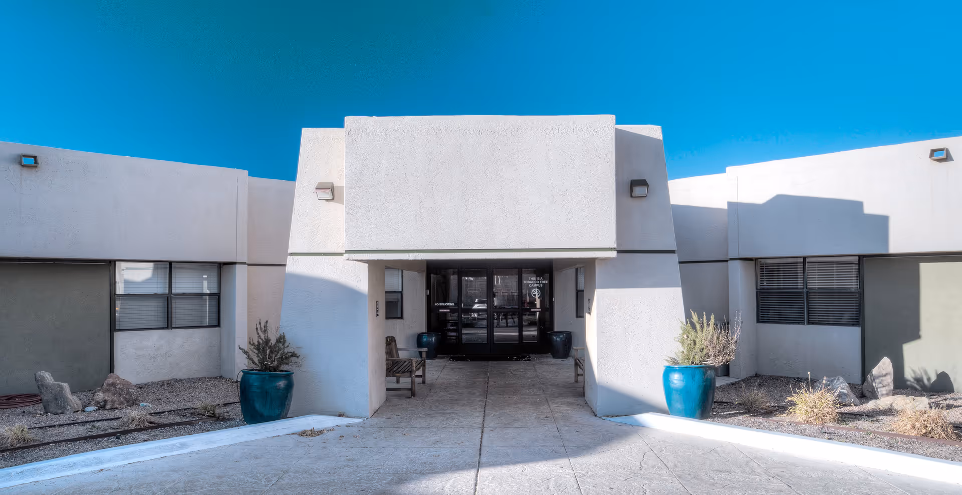 Front exterior view of Bear Canyon Rehabilitation Center building with a wide entrance, two large blue planters with plants on either side, and a clear blue sky above.