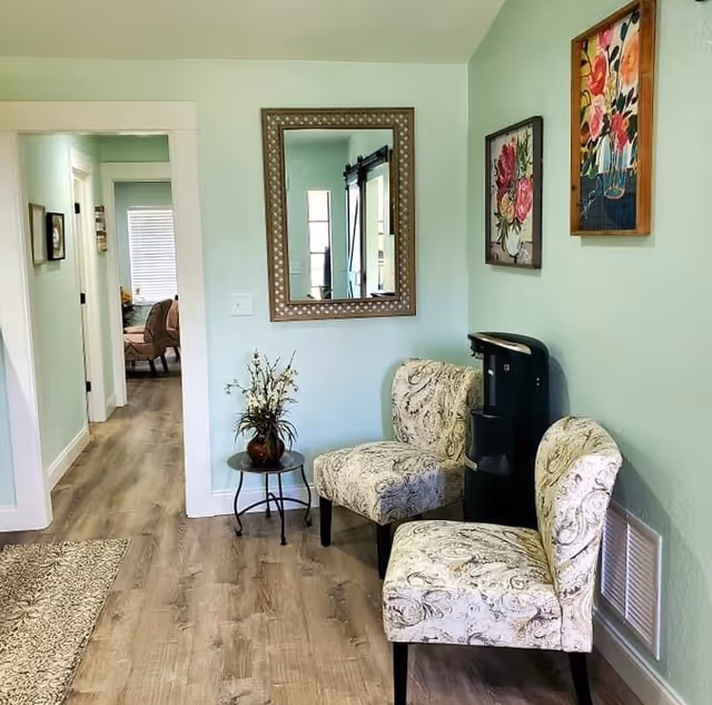 A cozy interior space with light green walls and wood flooring featuring two patterned upholstered chairs, a small round table with a flower arrangement, a water dispenser, and two floral paintings on the wall. A large decorative mirror hangs on the wall, reflecting part of the room and a hallway leading to another room with a window and additional seating.