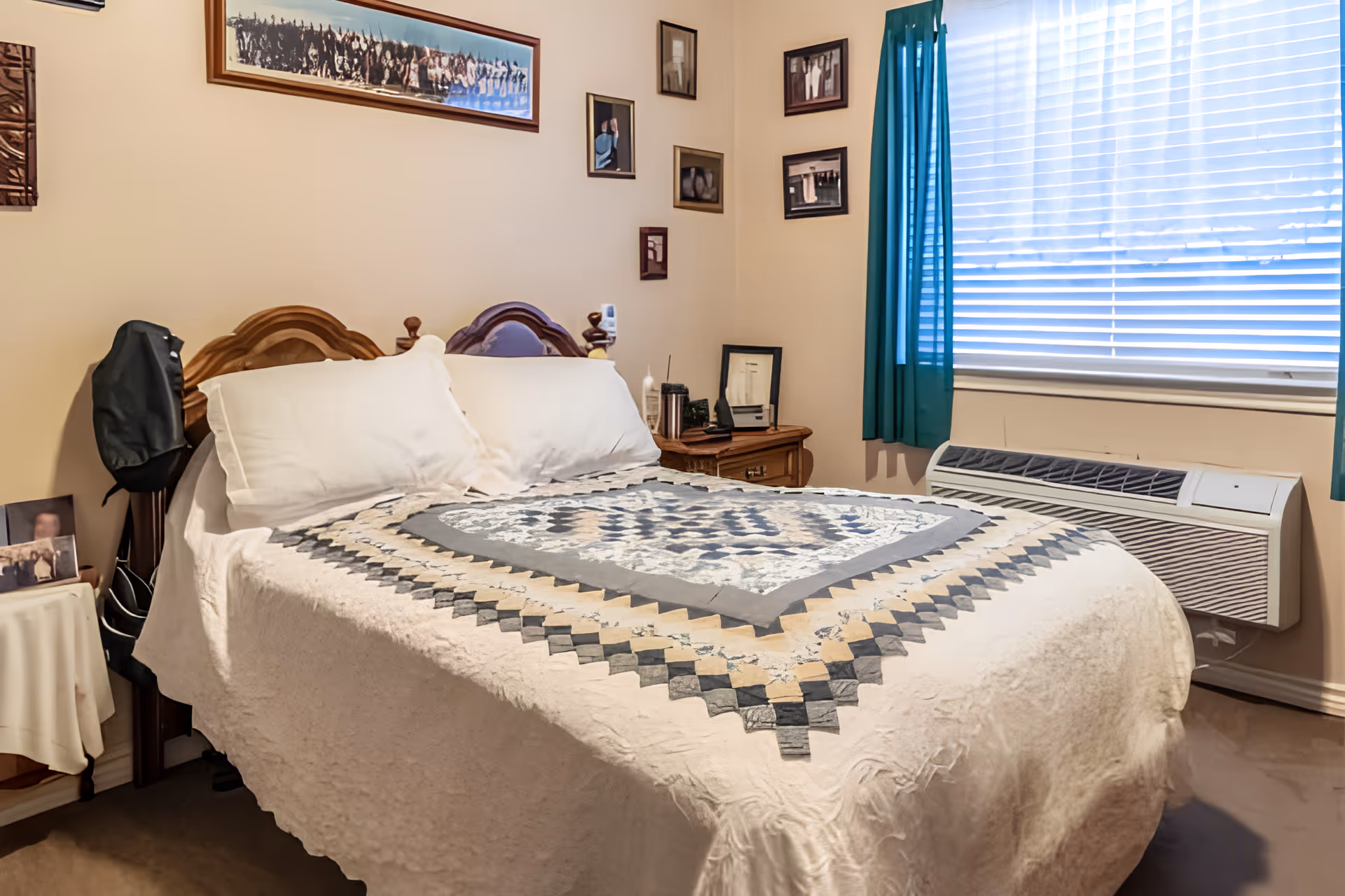 A cozy bedroom with a double bed covered by a quilted blanket in shades of gray, beige, and white. The bed has two white pillows and a wooden headboard. On the right side, there is a window with white blinds and teal curtains, and below it, a wall-mounted air conditioning unit. The walls are decorated with several framed pictures and photographs. A wooden nightstand with various items is next to the bed.